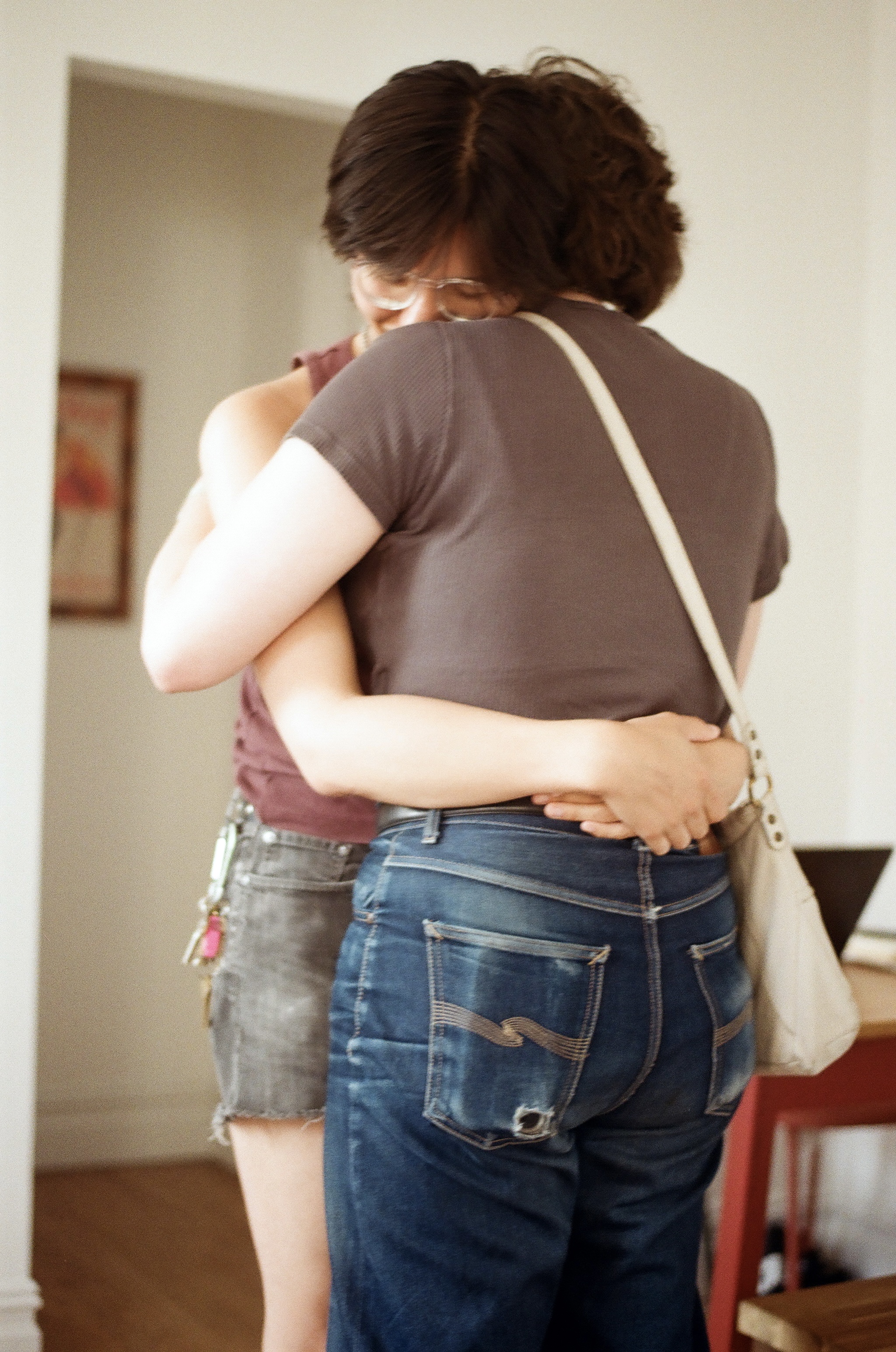 Two lesbians embracing. Neither are facing the camera, but laying their heads on each other's shoulders. In the foreground, one wears faded jeans with a hole in the back pocket. In the background, the other wears shorts with a carabiner.