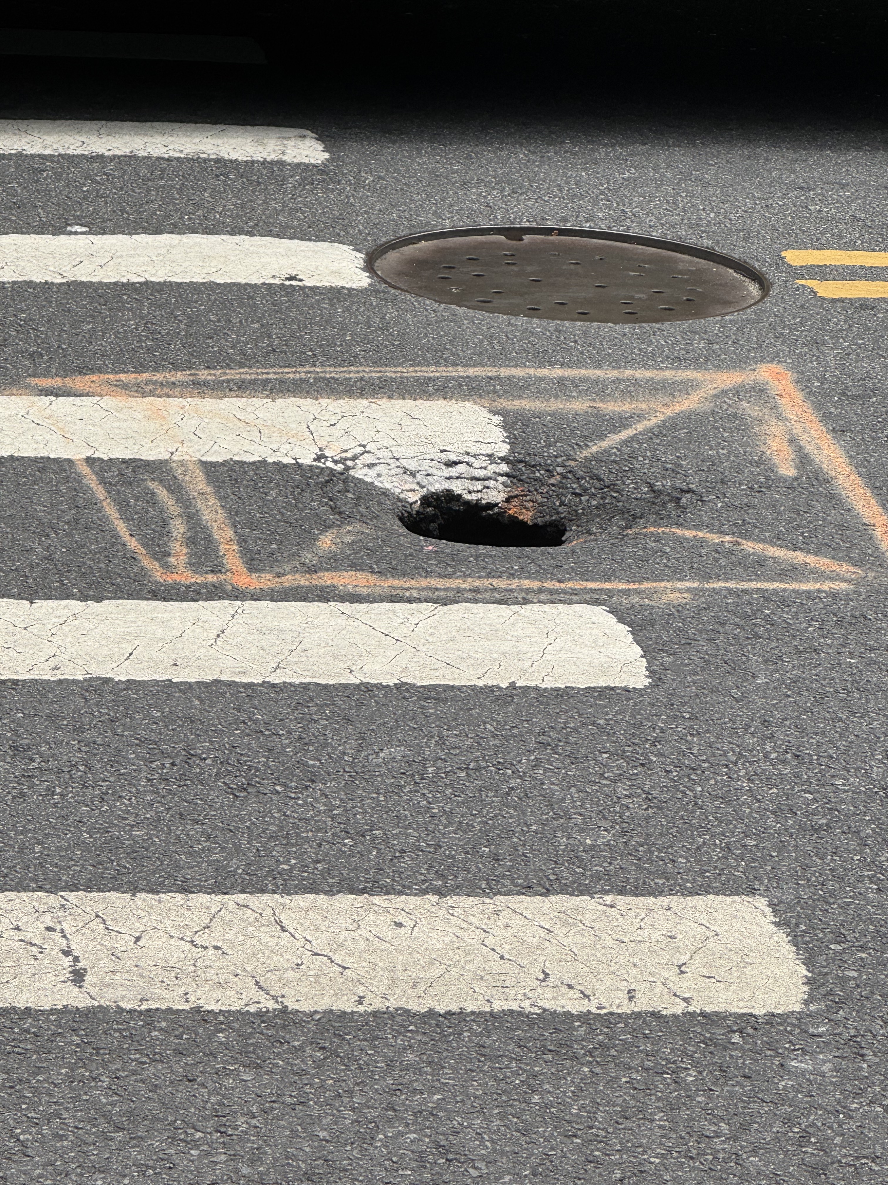 A small sinkhole in the middle of a crosswalk. A covered manhole is behind the sinkhole. The sinkhole is marked with chalk.